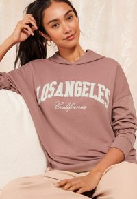 Mikina s kapucňou v púdrovej ružovej farbe s nápisom "LOS ANGELES California" vytlačeným na bielo. Mäkká tkanina, voľný strih, rebrované manžety a lem.