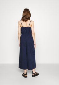 Navyblauer Jumpsuit mit Spaghetti-Trägern, elastischem Taillenbund und weitem Bein. Kombiniert mit schwarzen Sandalen mit strukturiertem Finish.