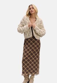 Gruba kremowa kardigan z warkoczowym splotem na brązowej, kratkowanej sukience maxi, zestawiona z beżowymi botkami za kolano. Teksturowane włókna i warstwowy design.