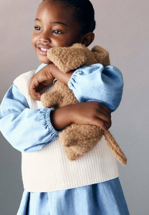 Ein Kind in einem hellblauen Shirt mit gepufften Ärmeln und einer cremefarbenen Strickweste umarmt ein braunes, flauschiges Stofftier mit Ohren und einem Schwanz.