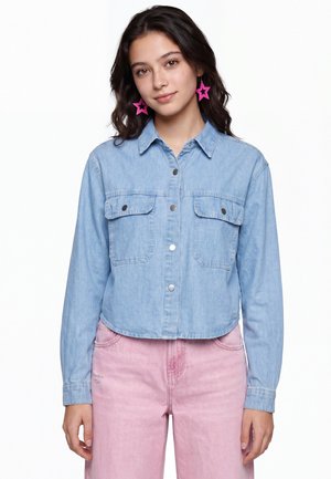 Giovane donna con capelli scuri che indossa orecchini rosa a forma di stella, una camicia di jeans azzurro chiaro con bottoni e pantaloni rosa su sfondo bianco.