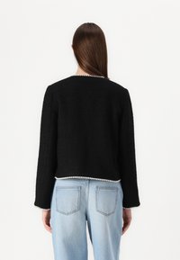 Veste noire texturée courte avec des manches longues et un accent en perles le long de l'ourlet, associée à un jean en denim bleu clair.