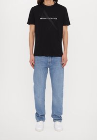 Svart bomullströja med "ARMANI EXCHANGE" i vit text och en stor logotyp, kombinerad med ljusblå denimjeans och vita sneakers.