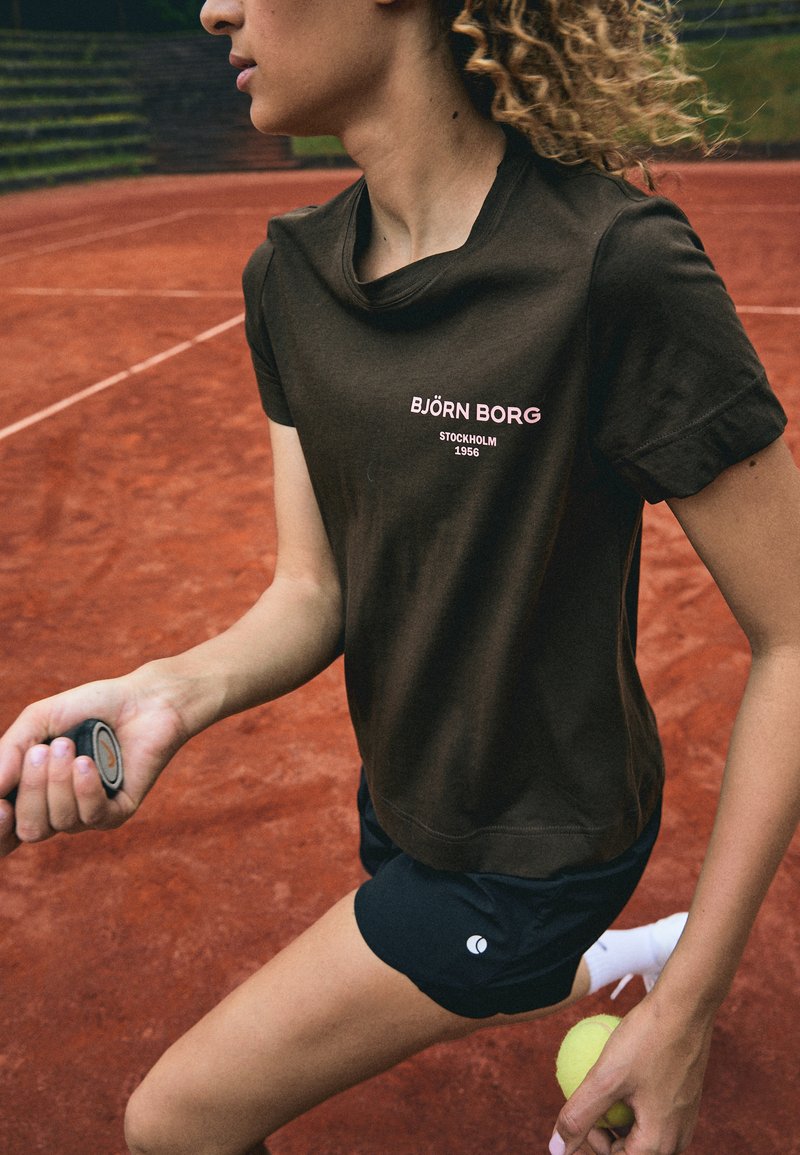 Maglietta marrone a maniche corte con scollo rotondo, caratterizzata da un logo rosa "BJÖRN BORG" sul petto, abbinata a pantaloni corti neri su un campo da tennis.