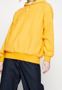 Keltainen fleecetakki, jossa on kiristysnauhalla varustettu huppu ja resorit helmassa. Edessä pieni valkoinen logo rinnassa. Yhdistetty tummien farkkujen kanssa.