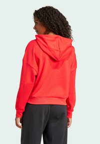 Röd hoodie tillverkad av mjukt material, med en stor huva och avslappnad passform med sänkta axlar. Bärs med svarta byxor, visas bakifrån.