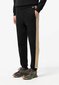 Černé tepláky s béžovým postranním pruhem, elastickým pasem a kapsami, doplněné zeleno-černými teniskami s červenými detaily.