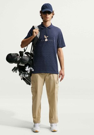 Jonge man die een marineblauw golfshirt, kaki cargobroek, pet en zonnebril draagt en een golftas met clubs over zijn schouder houdt.