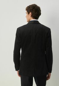 Mustat bleiseri, jossa on teksturoitu kuvio, slim fit -leikkaus, huippukaulus ja yksi halkio. Varustettu kahdella napilla ja hienovaraisella kiillolla, yhdistettynä mustiin housuihin.