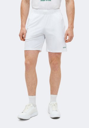 Άνδρας που φοράει λευκά ελαστικά σορτς μέσης με το λογότυπο «BORG» στο αριστερό πόδι, συνδυασμένα με λευκές κάλτσες και λευκά αθλητικά παπούτσια, στέκεται πάνω σε απλό φόντο.