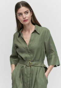 Sukienka koszulowa z lnu w oliwkowym kolorze, z kołnierzykiem, krótkimi rękawami i paskiem w talii z złotą okrągłą klamrą. Gładka tekstura i minimalistyczny design.