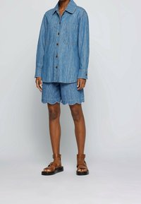 Ljusblå denimskjorta med två bröstfickor, knäppt, matchad med shorts med vågigt avslut; modellen bär bruna sandaler med ankelsnören.