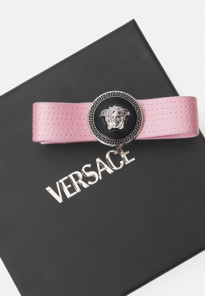 Rozā satīna josta ar melnu apaļu sprādzi, kas rotāta ar sudraba Medūzas simbolu un grieķu atslēgas motīvu, novietota uz melnas Versace kastes.