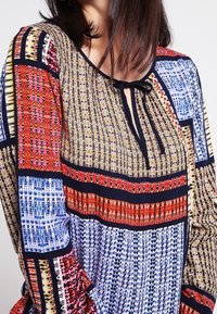 Blusa multicolore con un diverso motivo geometrico in rosso, blu e giallo. Tessuto morbido, scollatura rotonda con un fiocco e maniche lunghe.