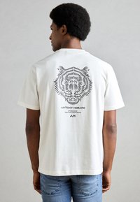 T-shirt in cotone crema con una grafica di una tigre nera sul retro, con il nome del marchio "ANTONY MORATO" e l'anno di fondazione stampati sotto.