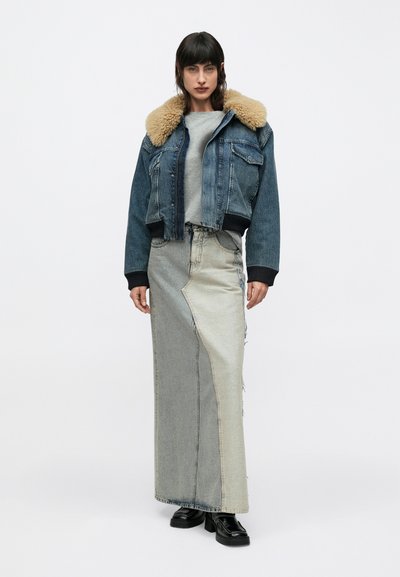 Moteris dėvi džinsinę striukę su smėlio spalvos kailio apykakle, pilką marškinėlį, ilgą džinsinę sijoną su lopais ir juodus platforminius batus, stovėdama prieš baltą foną.