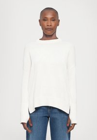 Pull en tricot blanc avec ourlet et poignets côtelés, présentant une coupe décontractée et un léger design haut-bas. Détail de logo subtil sur le devant.