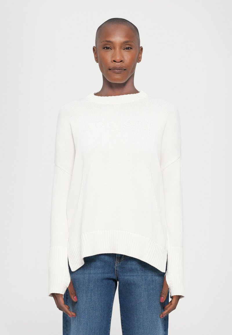 Pull en tricot blanc avec ourlet et poignets côtelés, présentant une coupe décontractée et un léger design haut-bas. Détail de logo subtil sur le devant.