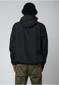 Chaqueta impermeable negra con capucha, que presenta una textura suave y puños elásticos, combinada con pantalones verde oliva. Sombrero de punto negro en la cabeza.