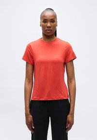 Γυναίκα με ίσια μαλλιά μαζεμένα πίσω, που φοράει απλό κόκκινο κοντομάνικο πουκάμισο και μαύρο παντελόνι, στέκεται μπροστά σε λευκό φόντο.