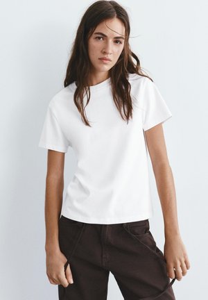 Mladá žena s dlouhými hnědými vlasy, oblečená v jednoduchém bílém tričku s krátkými rukávy a tmavých kalhotách, stojící před světle šedým pozadím.