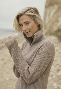 Vrouw met kort blond haar in een beige gebreide zip-up trui, staand op een rotsachtig strand met kliffen en zee op de achtergrond.