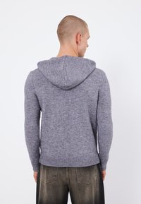 Grauer gestrickter Kapuzenpullover mit langen Ärmeln, strukturiertem Stoff, geripptem Saum und Kordelzug-Kapuze, von hinten betrachtet, kombiniert mit dunklen Jeans.