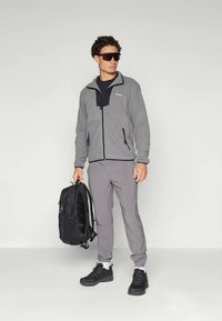 Grå fleecejakke med svarte detaljer, grå bukser og svart ryggsekk. Modellen har på seg mørke solbriller og svarte joggesko. Jevn tekstur, tettsittende design.