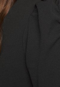 Svart texturerat tyg med en lätt lyster, med en oversized draperad ärmdekoration. Hår är delvis synligt i kompositionen.