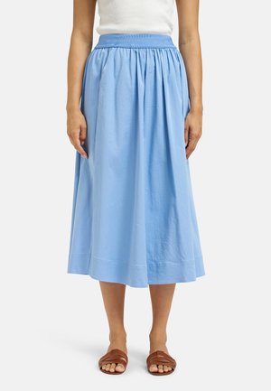 Frau trägt einen hellblauen Midirock mit geraffter Taille und ein weißes ärmelloses Oberteil, kombiniert mit braunen flachen Sandalen.