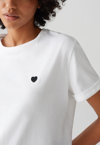 Weißes Baumwoll-T-Shirt mit umgeschlagenen kurzärmeln und einem kleinen schwarzen Herz, das auf der linken Brust gestickt ist. Glatte Textur, lässige Passform.