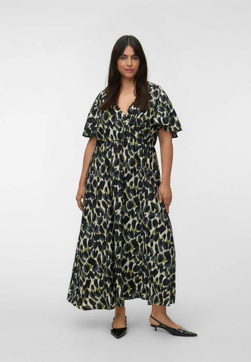 Femme aux longs cheveux foncés portant une robe maxi imprimé léopard noire, blanche et verte et des chaussures slingback noires à petit talon, debout.
