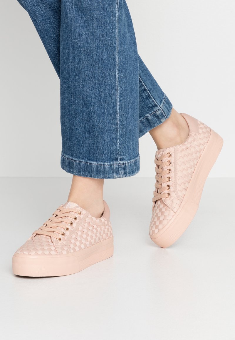 Pieds portant des baskets rose pâle à motif tissé et un jean denim bleu court, debout sur un sol blanc.