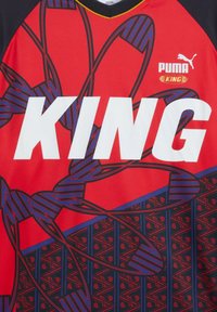 Maillot de sport rouge avec motifs abstraits noirs et bleus, grand texte blanc "KING" et logo Puma blanc avec "KING" en or en dessous.