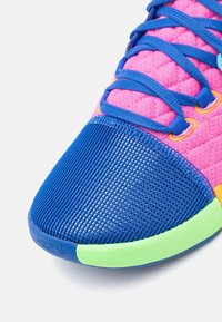 Gros plan sur la pointe d'une basket avec un mesh bleu, un tissu matelassé rose, des lacets bleu vif, des œillets jaunes et un bord de semelle vert clair.