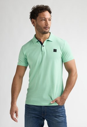 Mintgroen poloshirt van katoen, met een zwart merklabel, korte mouwen en een knoopsluiting met drie knopen. Draagt bij een blauwe spijkerbroek.