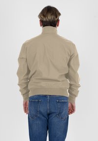 Giacca leggera beige con colletto alto, polsini e orlo elasticizzati, abbinata a jeans in denim blu. Texture liscia, design semplice.