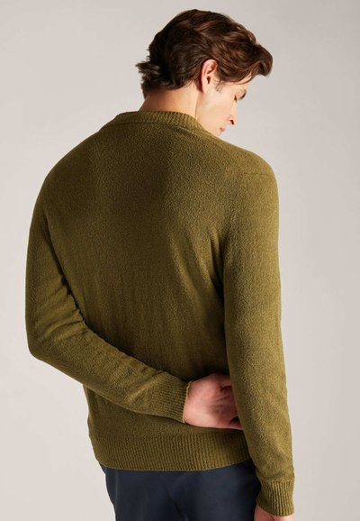 Una persona con cabello oscuro y ondulado lleva un suéter de punto verde, de espaldas, con una mano apoyada en la espalda. El fondo es neutro.