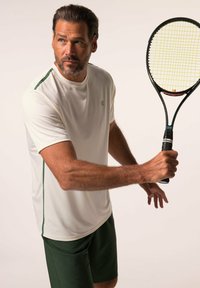 Camisa atlética blanca con acentos verdes, combinada con pantalones cortos verdes oscuros. La persona sostiene una raqueta de tenis, mostrando unos poderosos músculos en los brazos.