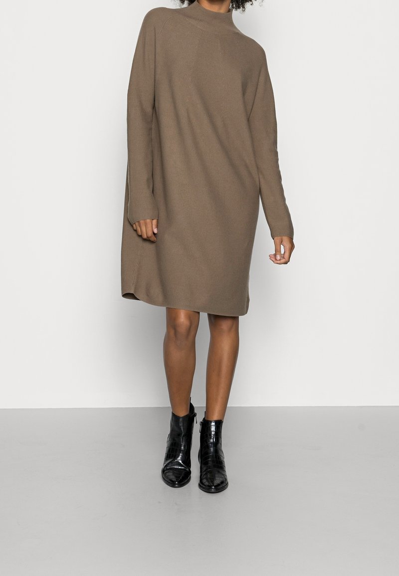 Robe tricotée marron avec des manches longues, un col montant et des fentes latérales. Portée avec des bottines noires à la cheville. Texture lisse et coupe décontractée.