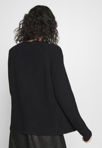 Personne avec des cheveux bouclés et foncés portant un pull en tricot noir texturé et un pantalon en cuir noir, de dos contre un fond clair.