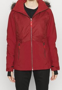Czerwona zimowa kurtka z kapturem wykończonym futrem, z przednim zamkiem błyskawicznym i kieszeniami na zamek, noszona na czarnej koszulce i czarnych spodniach.