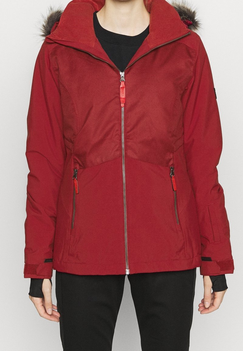 Czerwona zimowa kurtka z kapturem wykończonym futrem, z przednim zamkiem błyskawicznym i kieszeniami na zamek, noszona na czarnej koszulce i czarnych spodniach.