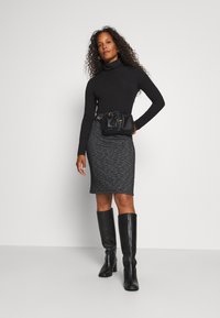 T-shirt à col roulé noir à manches longues, jupe crayon grise à motifs, bottes noires montantes et sac en bandoulière noir avec des accents dorés.
