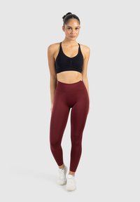 Zwarte cropped sportbeha met rimpeldetail, gecombineerd met hooggetailleerde kastanjebruine leggings. Model draagt witte sportschoenen. Neutraal grijze achtergrond.