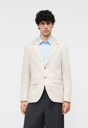 Νεαρός με μπούκλες φοράει ένα ανοιχτό μπεζ σακάκι πάνω από ένα ανοιχτό γαλάζιο πουκάμισο με κουμπιά και σκούρο, φαρδύ παντελόνι, στέκεται μπροστά σε λευκό φόντο.