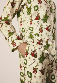 Pijamas de algodón con un patrón festivo que presenta al Grinch verde y renos, en crema con acentos rojos, verdes y amarillos. Textura a rayas visible.