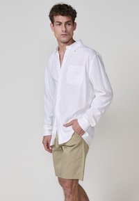 Cămașă albă cu mâneci lungi și un buzunar pe piept, purtată cu pantaloni scurți bej, având o croială relaxată și un material ușor texturat.