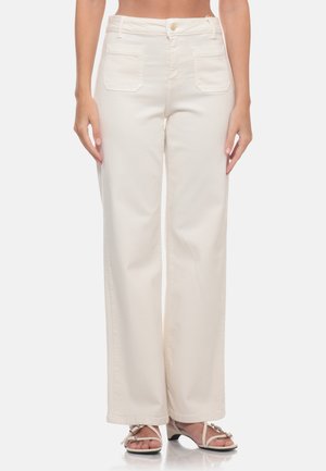 Donna che indossa pantaloni bianchi a vita alta e gamba larga con tasche applicate davanti e sandali beige con cinturini e tacco, in piedi su sfondo bianco.