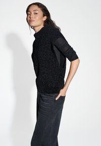 Maglione nero lavorato a maglia con collo alto e vestibilità ampia, abbinato a jeans in denim neri. Il maglione presenta un tessuto testurizzato e spacchetti laterali.